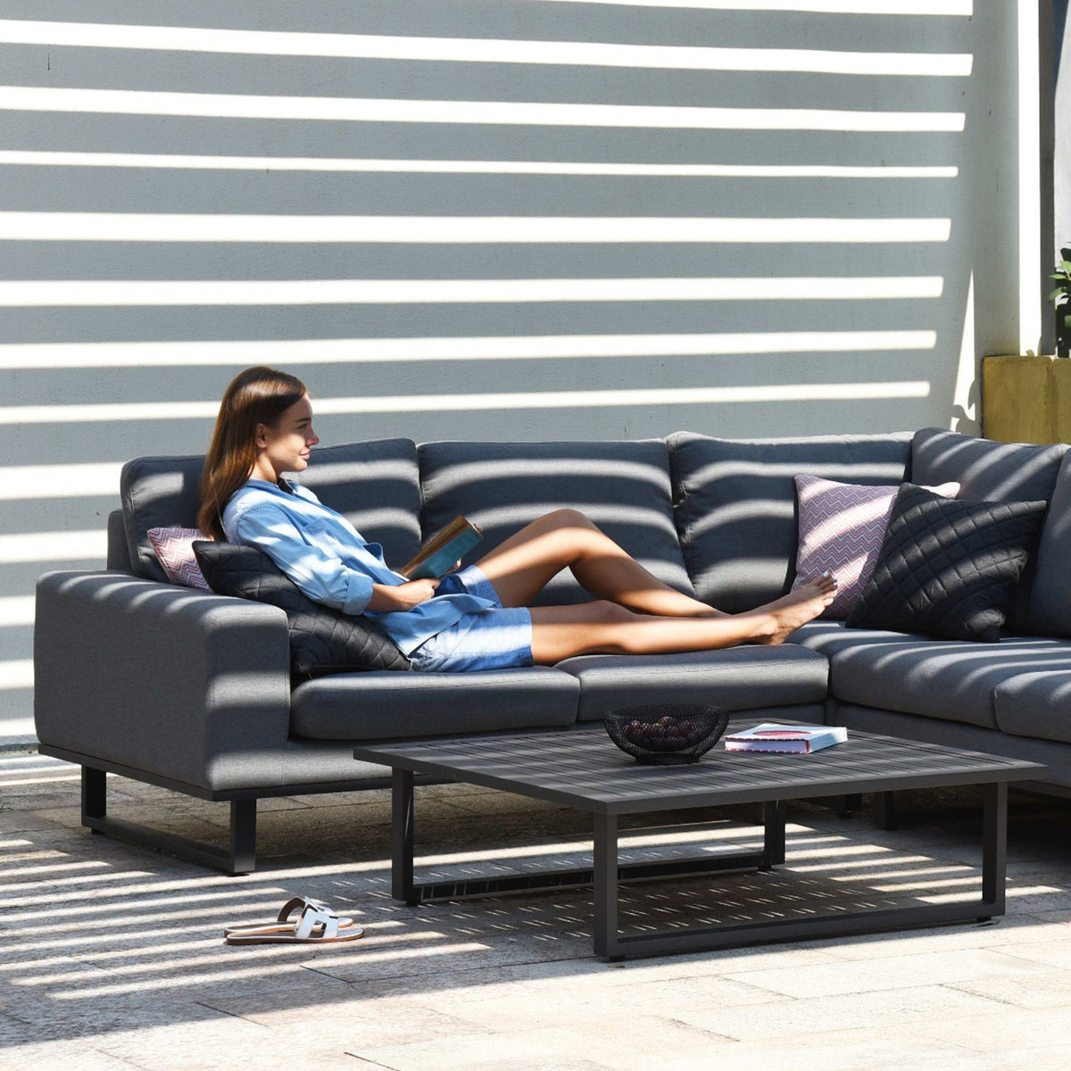 A person lounges on an outdoor sofa, reading a book in a sunny environment with striped shadows cast by slats overhead. Shoes and items rest on the ground beside a table.
