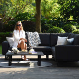 A person sits casually on an outdoor sofa, legs crossed, surrounded by lush greenery under bright sunlight, with a table in front of them holding a meal.