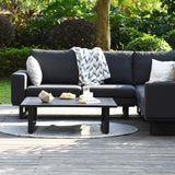 A black outdoor sofa with cushions and a throw, adjacent to a coffee table with snacks, is set on a patio surrounded by greenery.