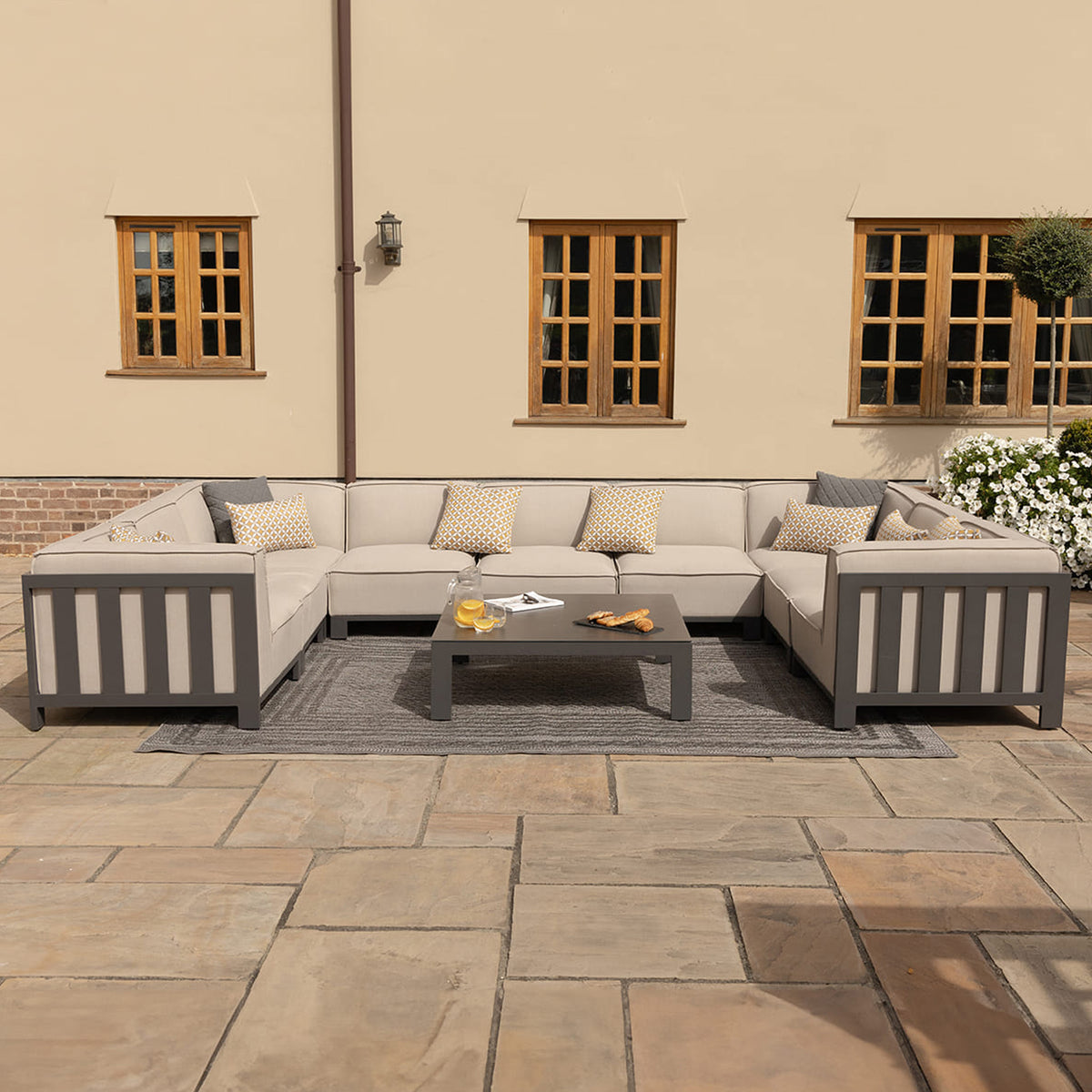 A sectional outdoor sofa and coffee table set on a patio, with cushions and decorative pillows, near a building with wooden windows and a lantern.