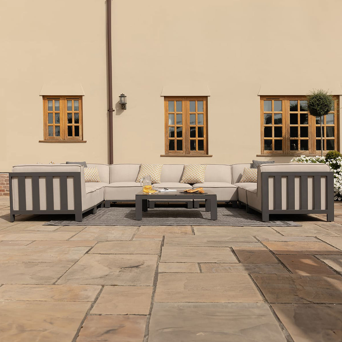 An outdoor sectional sofa is arranged in an L-shape on a patio, accompanied by a coffee table, with a background of a cream-colored wall with two windows.