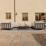 An outdoor sectional sofa is arranged in an L-shape on a patio, accompanied by a coffee table, with a background of a cream-colored wall with two windows.