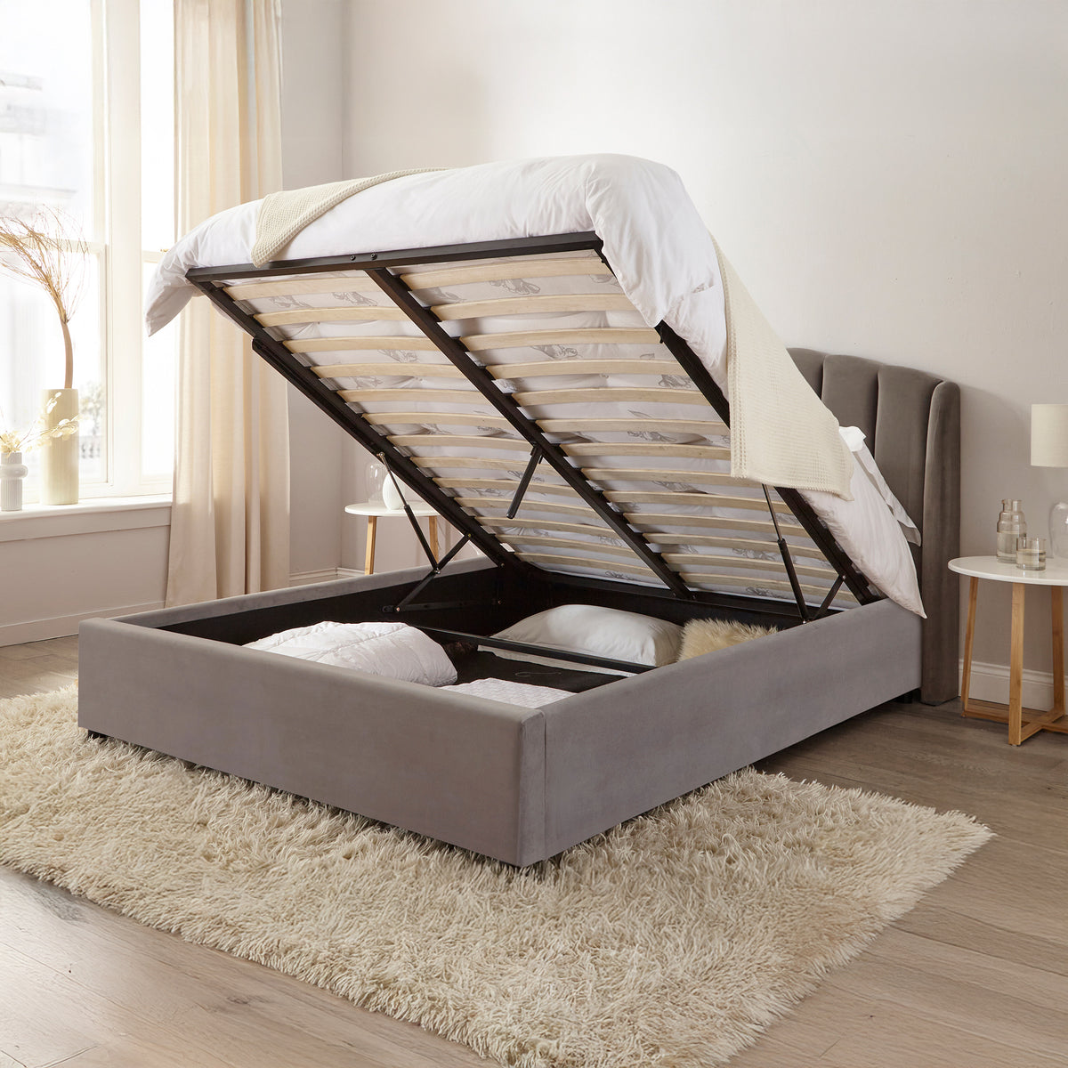 Gray upholstered bed with open storage compartments in a room with a window and side table.