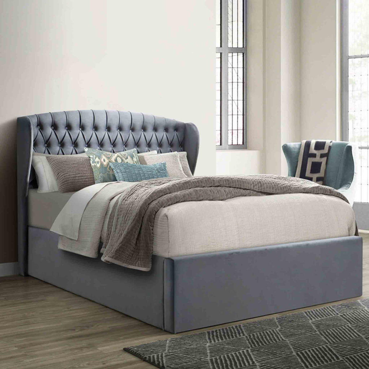A gray upholstered bed with a tall, tufted headboard is set against a paneled wall on herringbone-patterned wooden flooring.