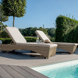 Two sun loungers by a pool with cushions, a hat, and a glass of water, set against a backdrop of trees and shrubs in a garden during daytime.