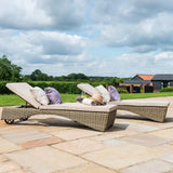 Two wicker sun loungers with beige cushions and pillows are set on a paved patio, under a clear blue sky with puffy clouds, surrounded by greenery.