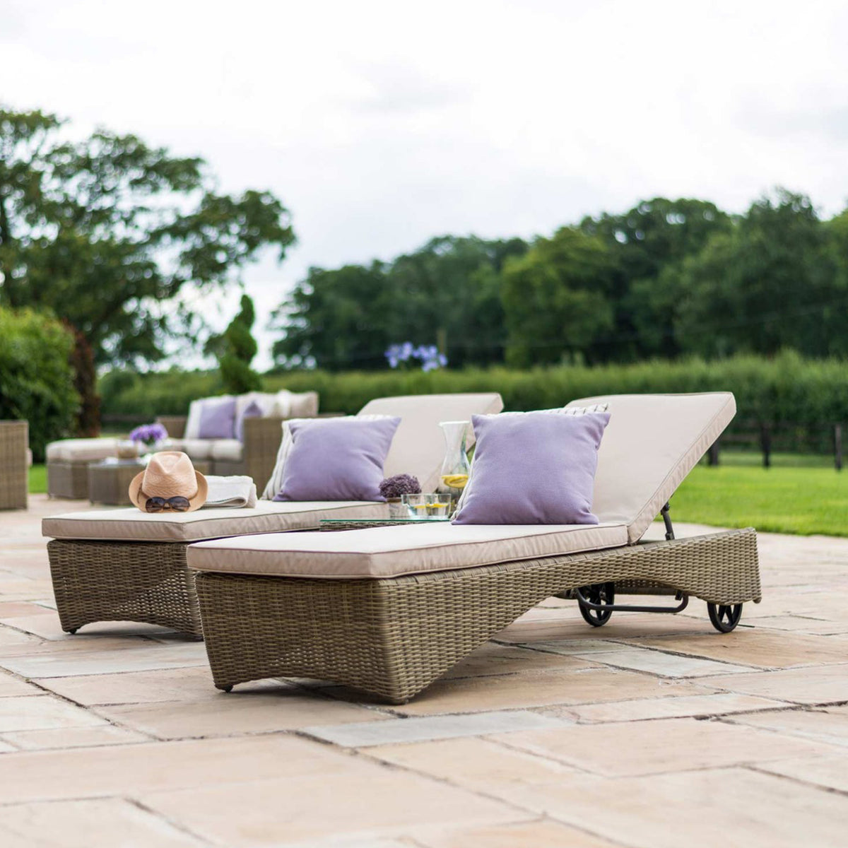A wicker sun lounger with cushions and a hat, set on a patio against a backdrop of lush greenery and a cloudy sky.