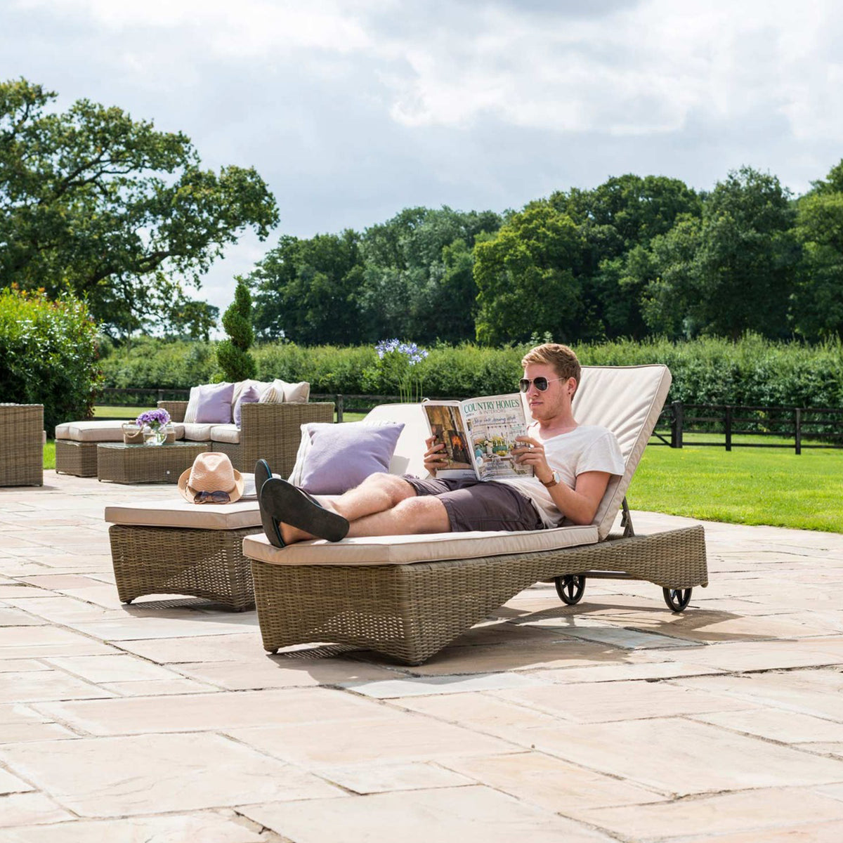 A person reclines on an outdoor sun lounger, reading a magazine titled "COUNTRY HOMES," amidst a patio setting with furniture and lush greenery in the background.