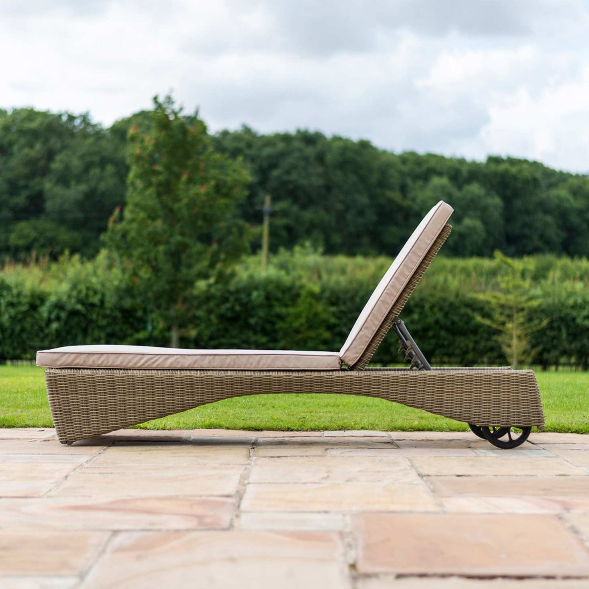 A wicker sun lounger with a beige cushion positioned on a paved patio, backed by a lush green lawn and trees.