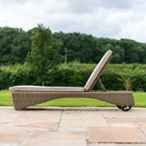 A wicker sun lounger with a beige cushion positioned on a paved patio, backed by a lush green lawn and trees.