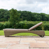 A wicker sun lounger with a cushion rests on a stone patio, amidst a lush green lawn and trees in the background.