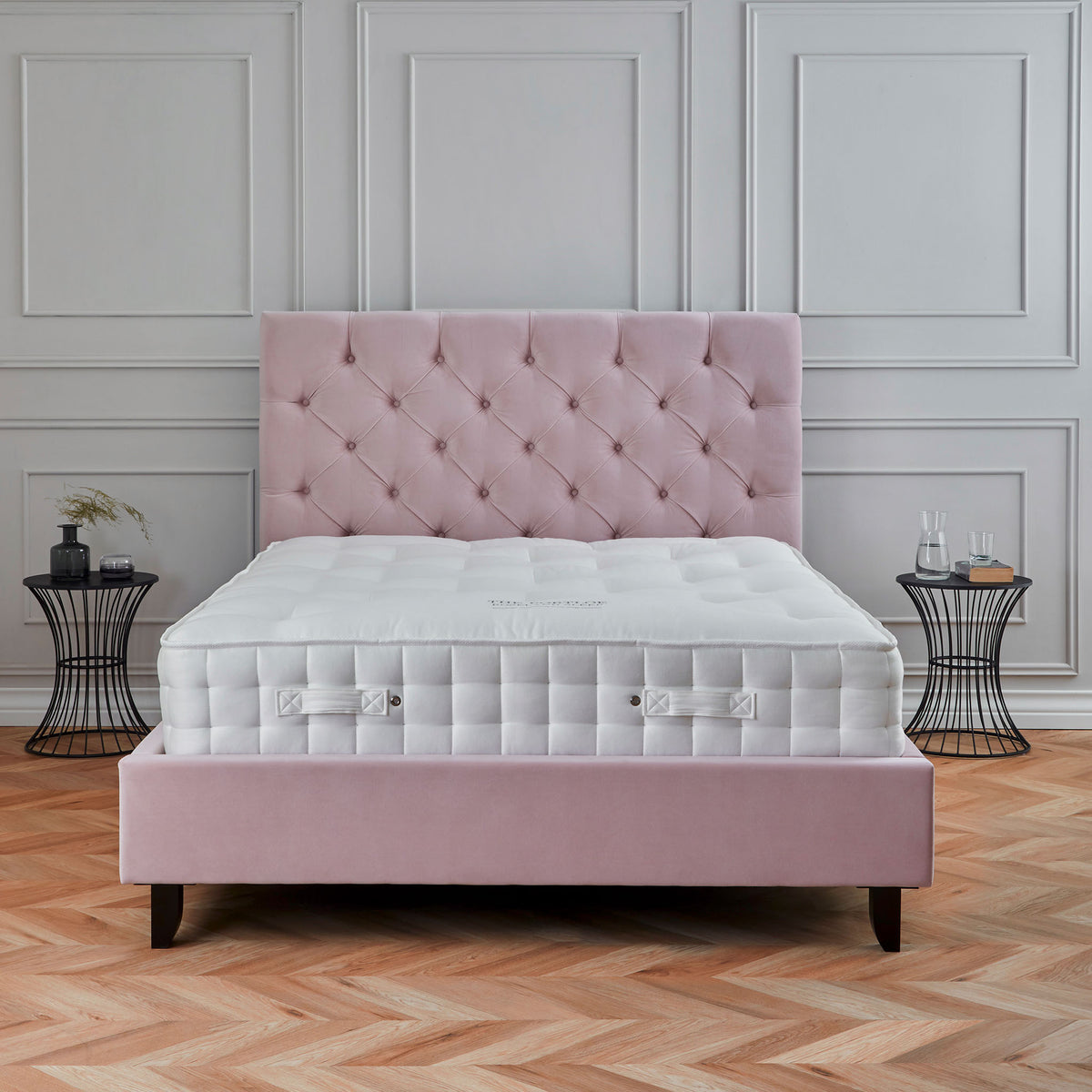 A plush pink bed with a tufted headboard sits in a room with classic paneled walls and a herringbone parquet floor. A small side table and decorative items accompany it.