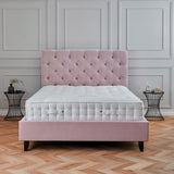 A plush pink bed with a tufted headboard sits in a room with classic paneled walls and a herringbone parquet floor. A small side table and decorative items accompany it.