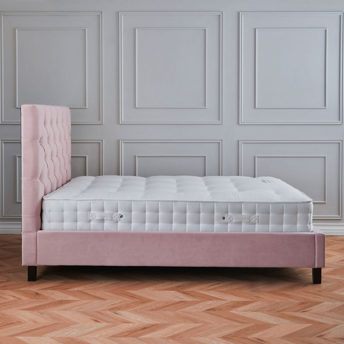 A pink, tufted single bed against a grey paneled wall, on a herringbone patterned wooden floor.