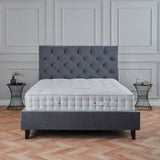 A gray upholstered bed with a quilted headboard and mattress set in a room with paneled walls and herringbone wood flooring, accompanied by a wireframe side table.