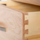 A close-up of a wooden drawer corner showing intricate dovetail joinery, against a blurred interior background.