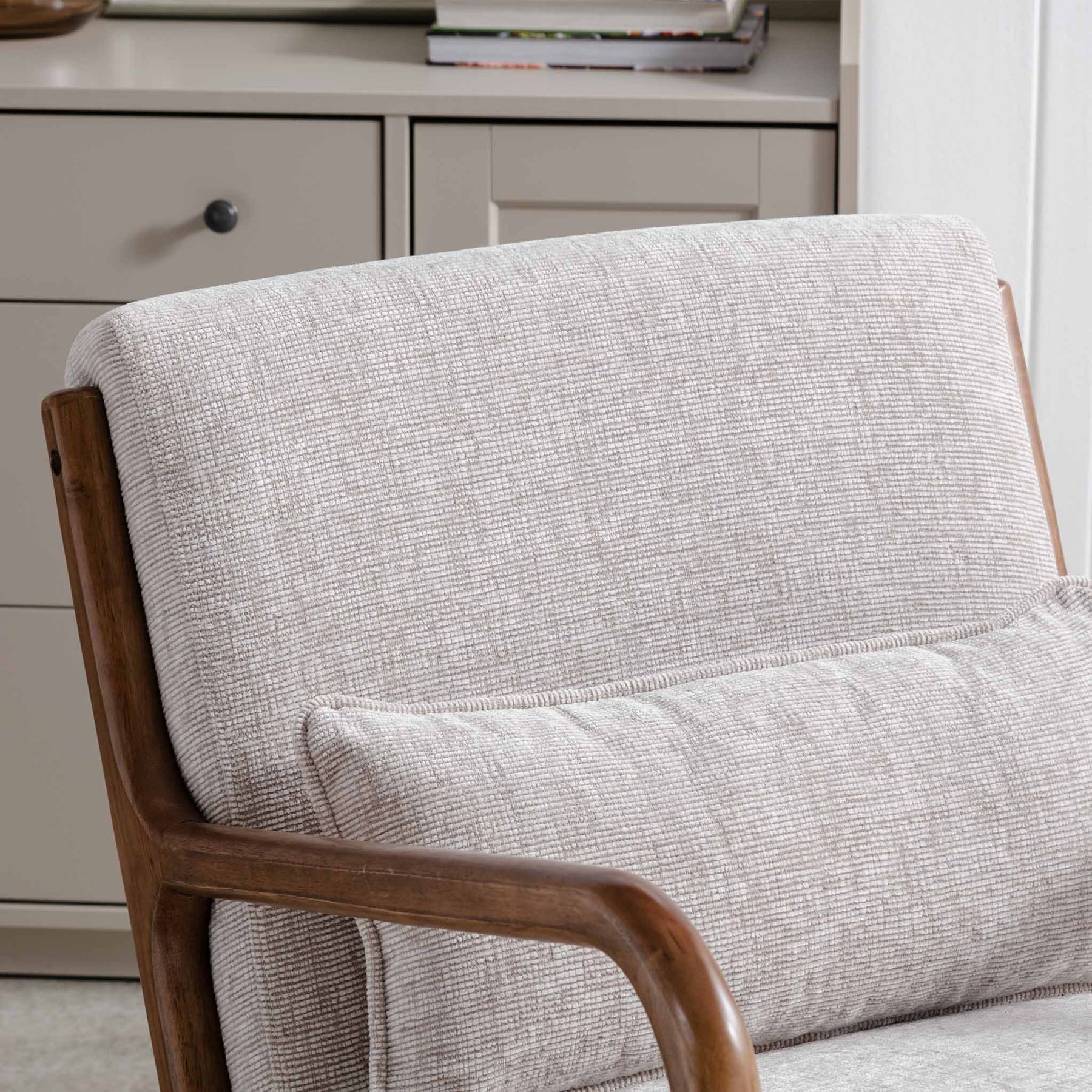 A beige upholstered chair with a wooden frame, positioned against a gray carpet, near a drawer unit with metal handles.