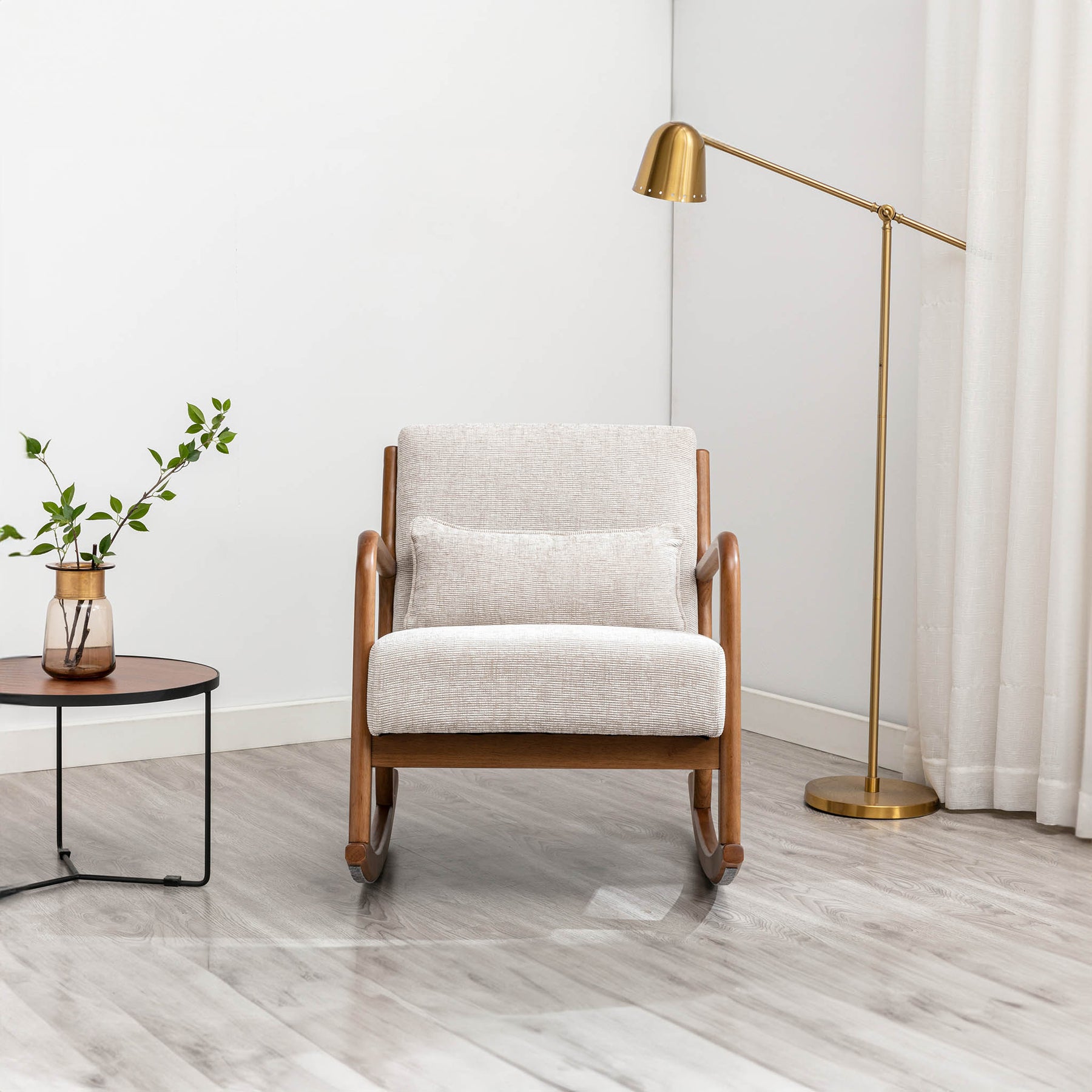 A beige armchair stands between a round side table with a vase and plant and a brass floor lamp, set against a white wall on a light wood floor.