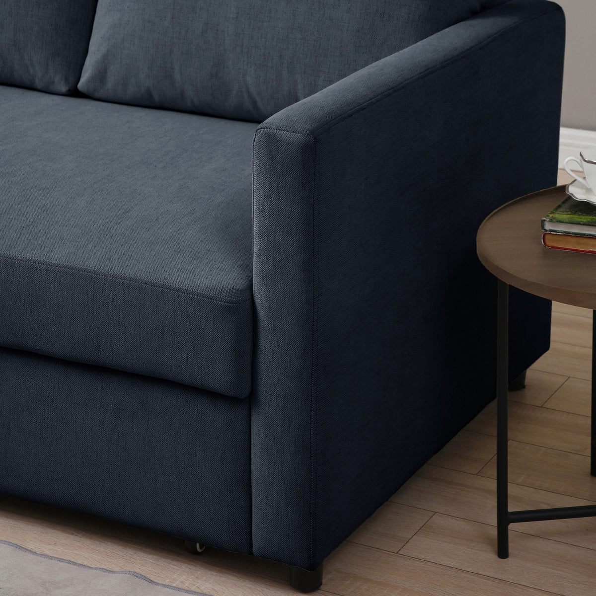 A navy blue sofa with a smooth texture, adjacent to a round wooden side table with books, placed in a room with wooden flooring.