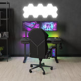 A gaming chair is positioned in front of a desk with dual monitors, a backlit keyboard, and headphones, set in a room with hexagonal wall lights and a modern aesthetic.