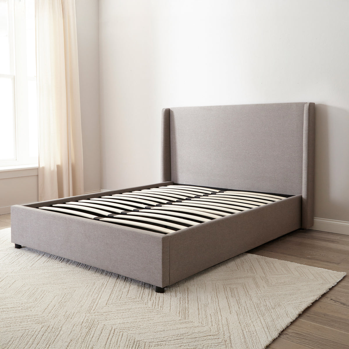 Brent Ottoman Storage Bed Frame from Roseland Furniture- Gray upholstered bed in a minimalistic bedroom setting with light-colored walls and wooden flooring.