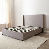 Brent Ottoman Storage Bed Frame from Roseland Furniture- Gray upholstered bed in a minimalistic bedroom setting with light-colored walls and wooden flooring.