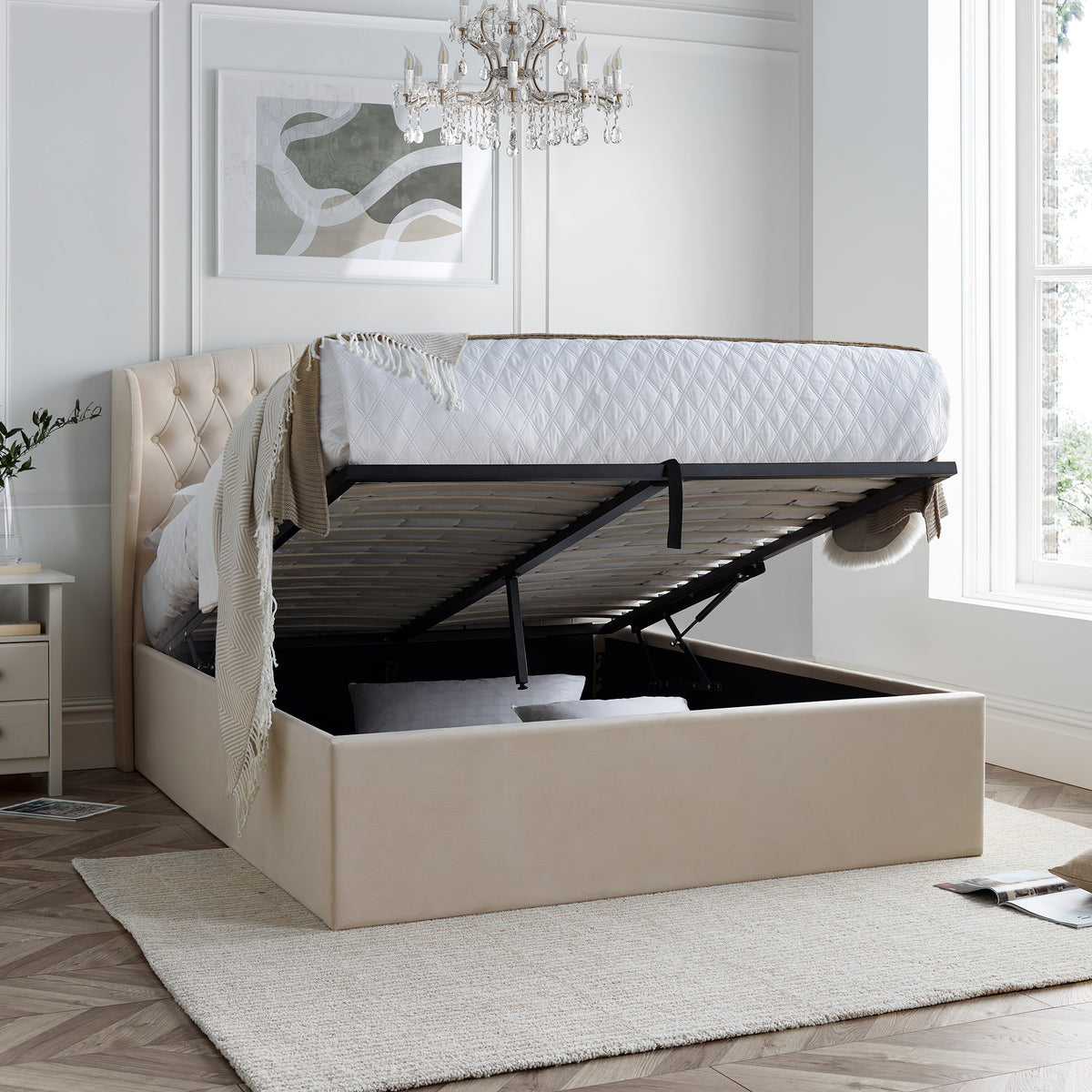 A natural upholstered ottoman bed with a lifted mattress reveals storage underneath, positioned against a paneled wall on a herringbone wood floor.