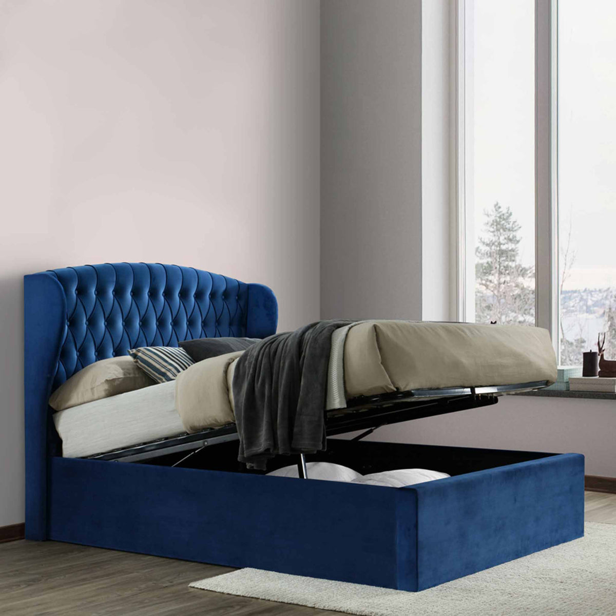 A blue upholstered storage bed with its mattress lifted to reveal an empty compartment underneath. It's situated in a room with paneled walls and herringbone wood flooring.