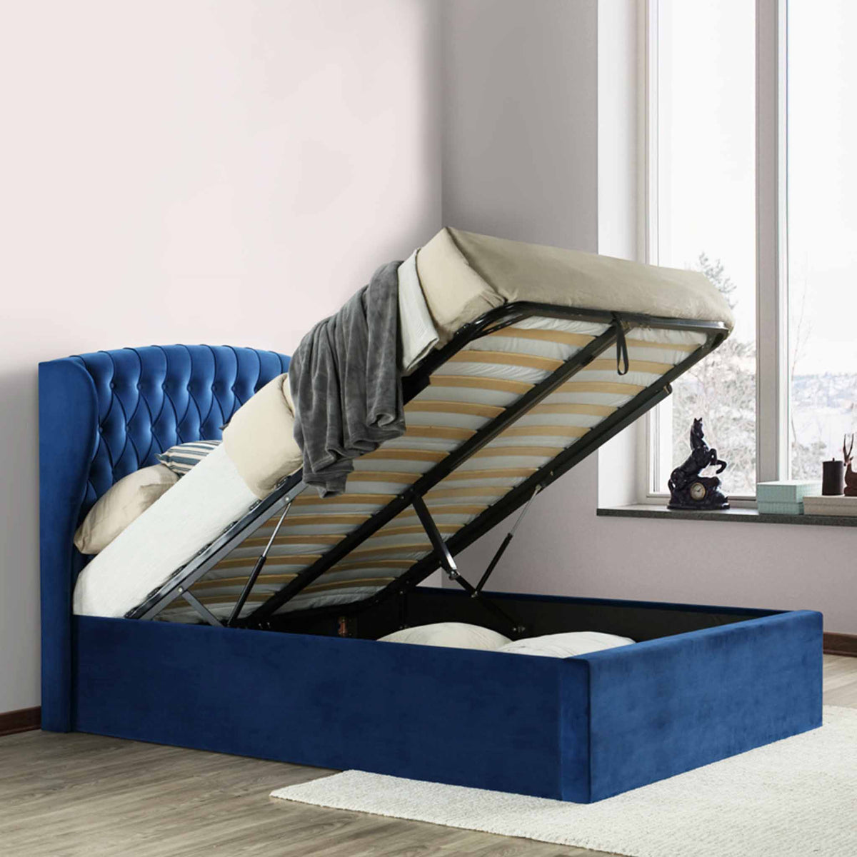 A blue upholstered storage bed with the mattress lifted up, revealing an open storage space beneath, set against a paneled wall on a herringbone floor.