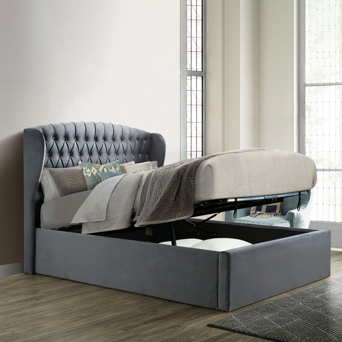 An open gray storage bed with a lifted mattress reveals a spacious compartment underneath, situated against a paneled wall on a herringbone-patterned wooden floor.
