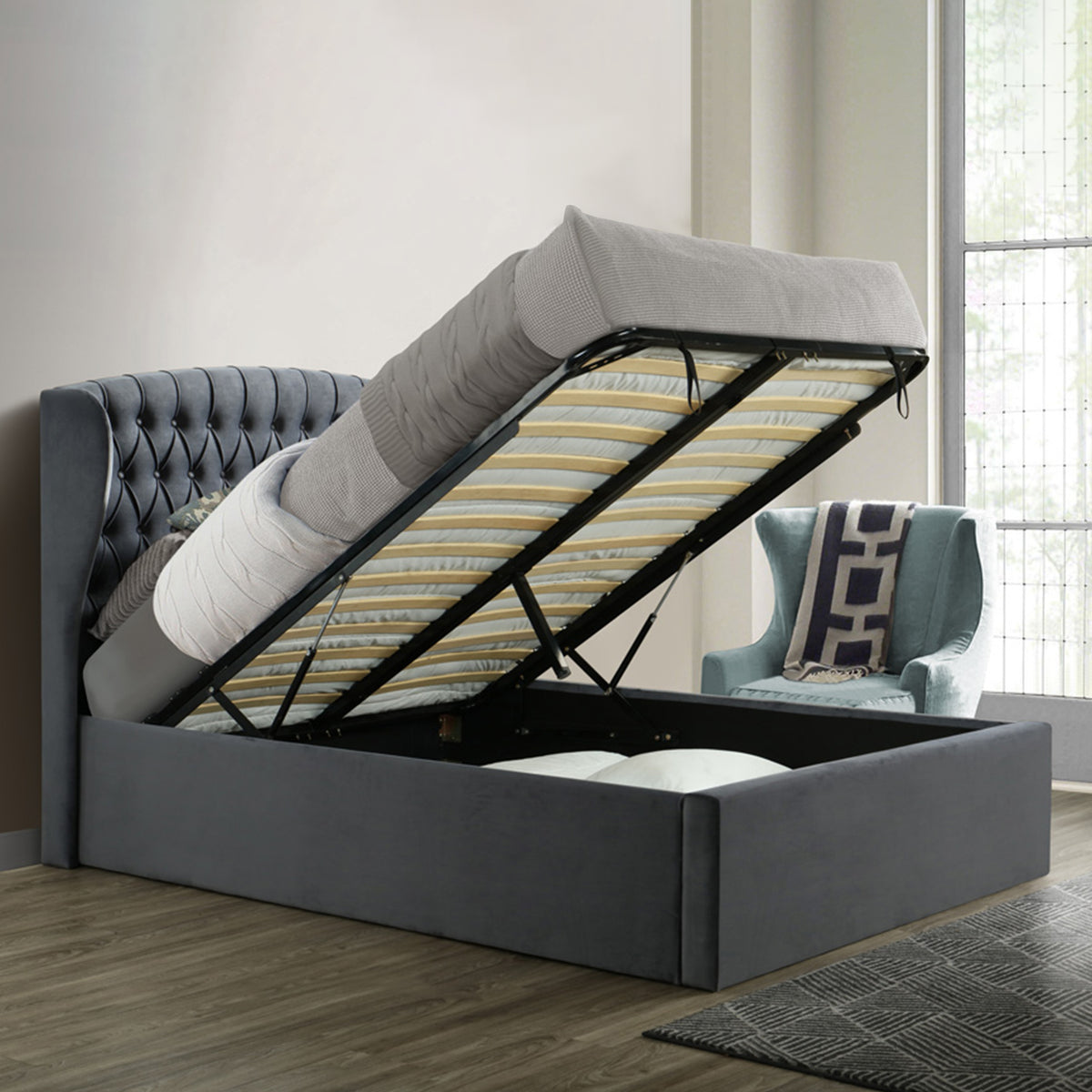 A gray upholstered bed with a lifted mattress reveals storage underneath, positioned against a paneled wall on a herringbone wood floor.