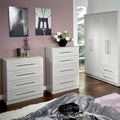 White bedroom furniture, including a chest of drawers and a wardrobe, is arranged in a room with lavender walls, dark flooring, and decorative items like wall art and a flower vase.