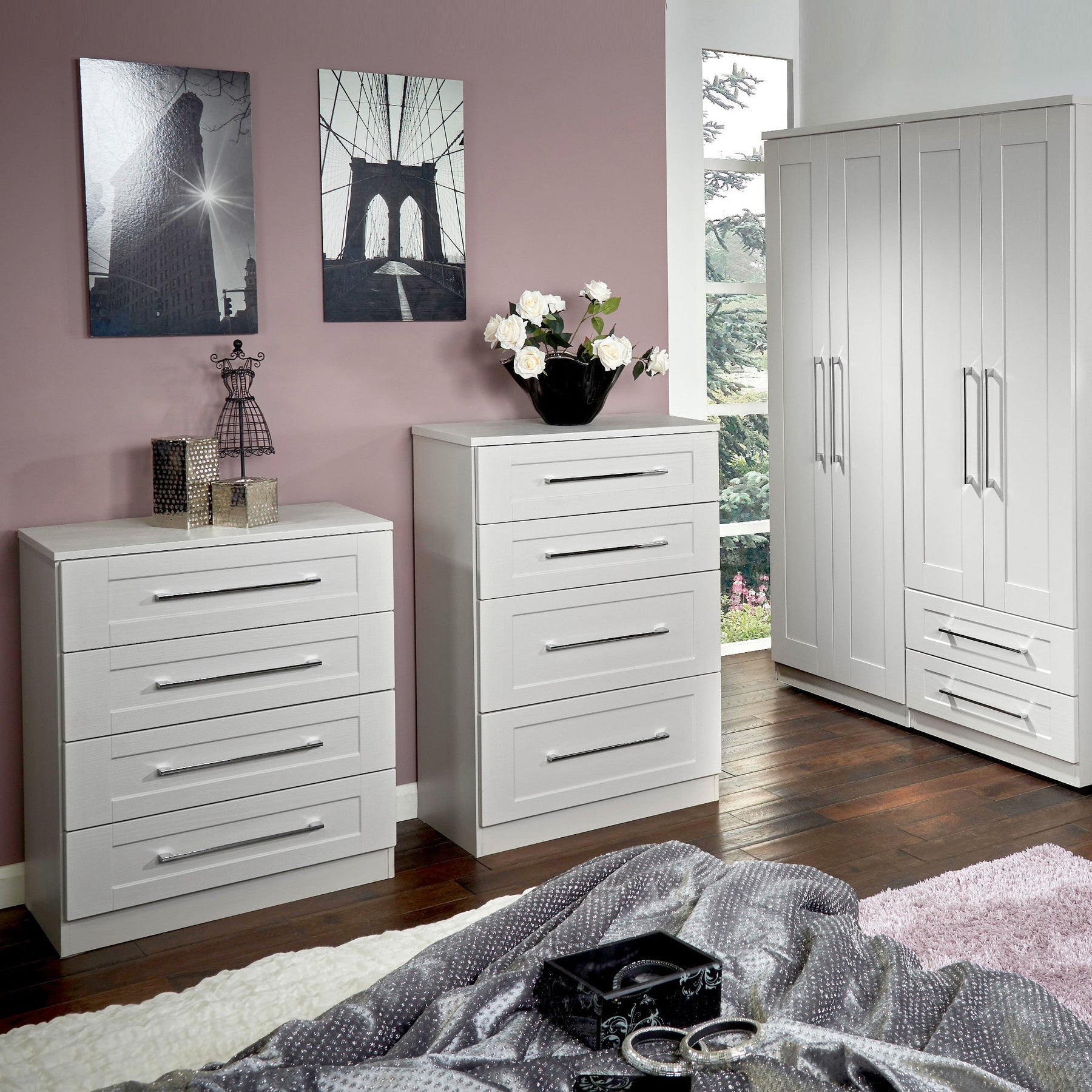 White bedroom furniture, including a chest of drawers and a wardrobe, is arranged in a room with lavender walls, dark flooring, and decorative items like wall art and a flower vase.