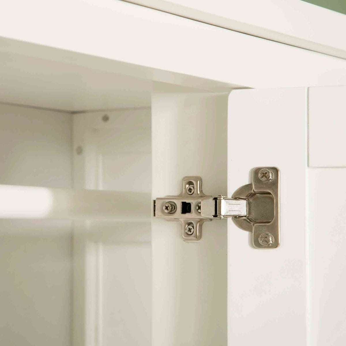 A metal cabinet hinge connects a white cabinet door to its frame, showing a close-up of its mechanism against a blurred interior background.