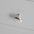 A metal drawer knob, affixed to a white surface, casting a soft shadow below, against a plain, neutral background.