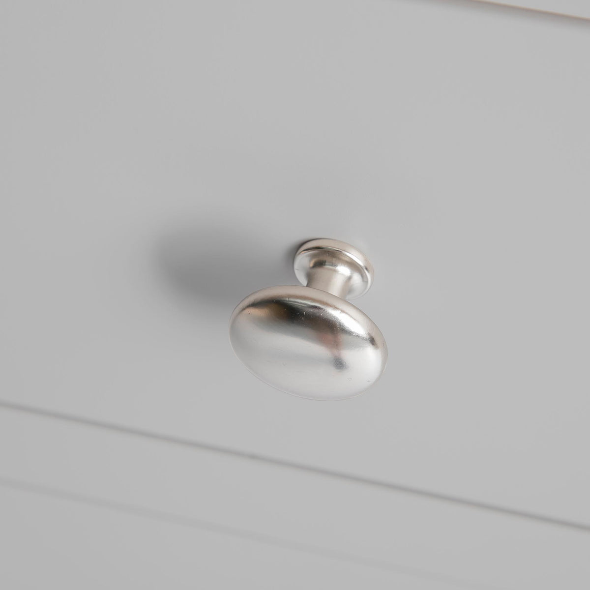 A metal drawer knob, affixed to a white surface, casting a soft shadow below, against a plain, neutral background.
