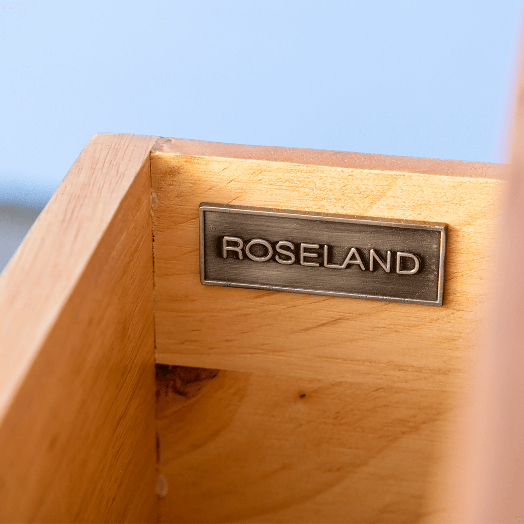 A metal plaque with the inscription "ROSELAND" is mounted on a wooden surface. The background is blurred with a hint of blue sky.