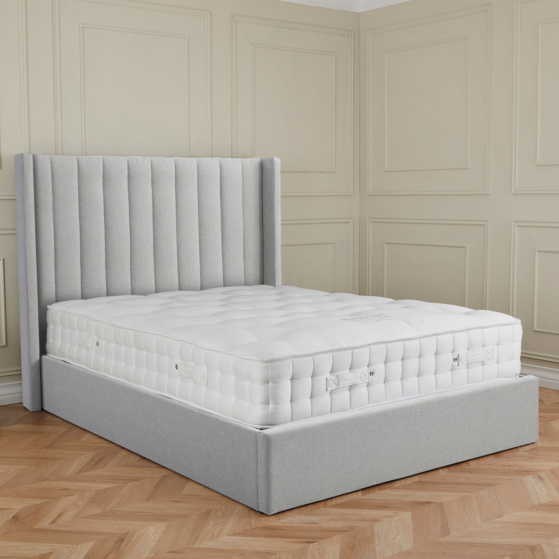 A grey upholstered bed with a plush mattress in an elegant room with paneled walls and herringbone wood flooring.