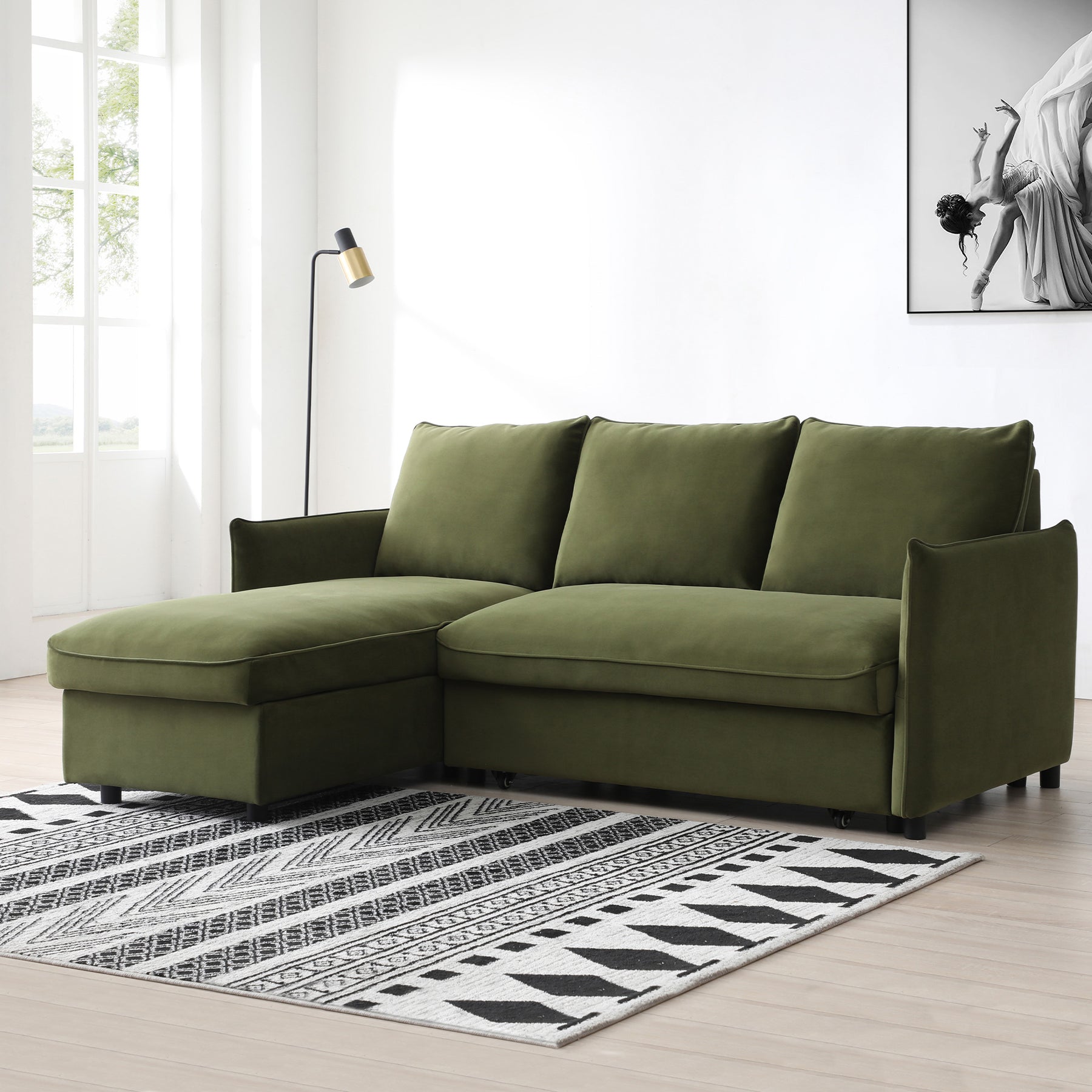 A green sectional sofa with chaise lounge sits on a patterned rug, under a floor lamp, beside a large window, with an art frame on the wall.