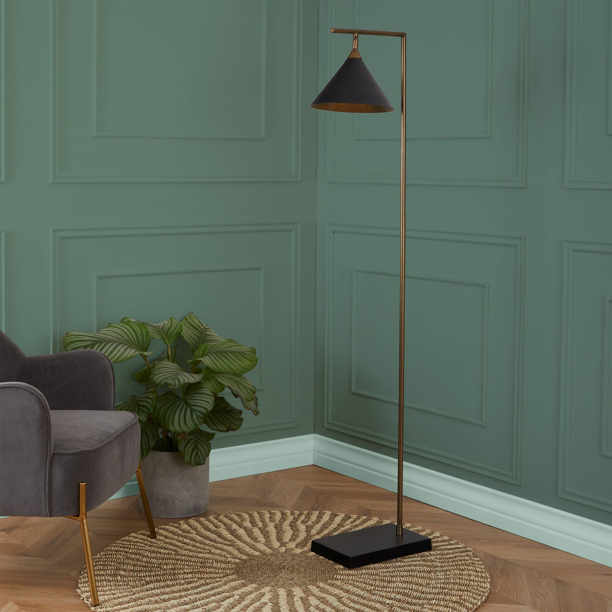 A black-shaded floor lamp with a brass stand illuminates a corner beside a grey armchair, with a green potted plant on a round woven rug against a teal wall.