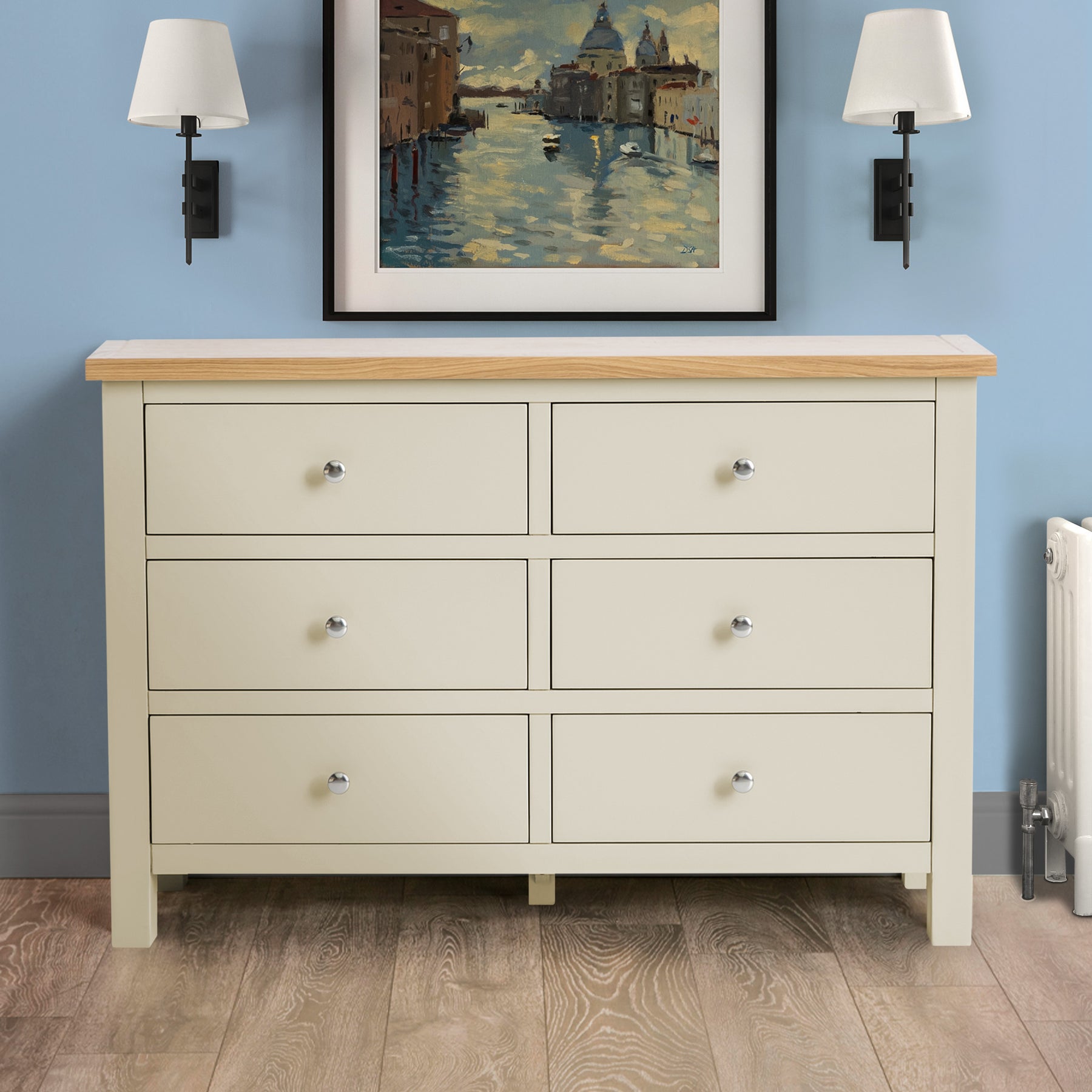 A cream-colored, six-drawer wooden dresser stands against a blue wall in a room with wood floors, flanked by wall lamps and beneath a framed painting.