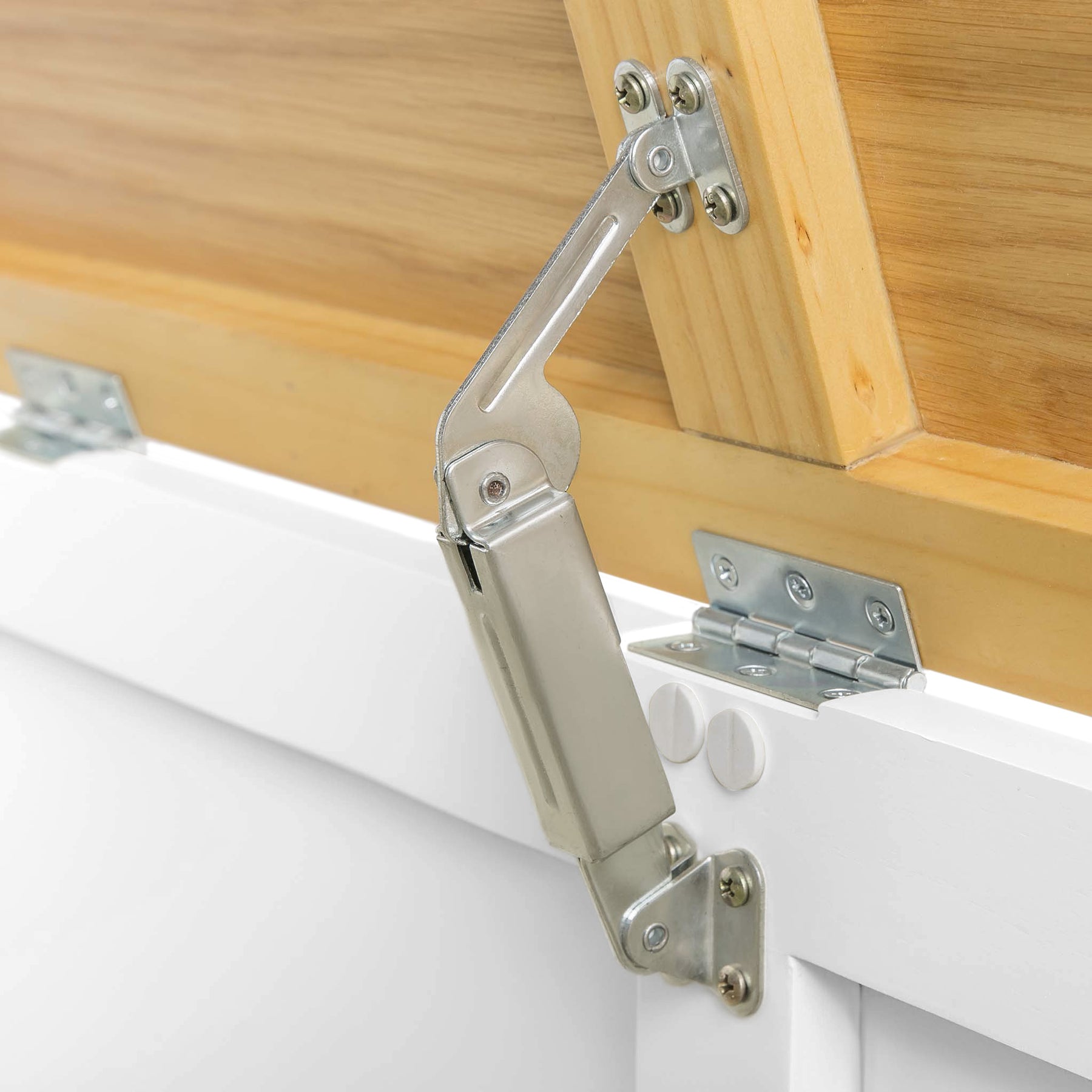 A silver gas spring door lift mechanism connects a wooden cabinet to its white door, holding it open against a plain background.