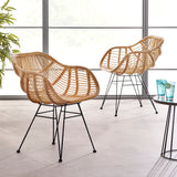 Two tan wicker chairs with black metal legs are placed on a light floor near a window, next to a small round table with two blue glasses.