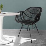 A black, woven wire chair sits next to a white round table with a plant and books, illuminated by sunlight against a teal wall.