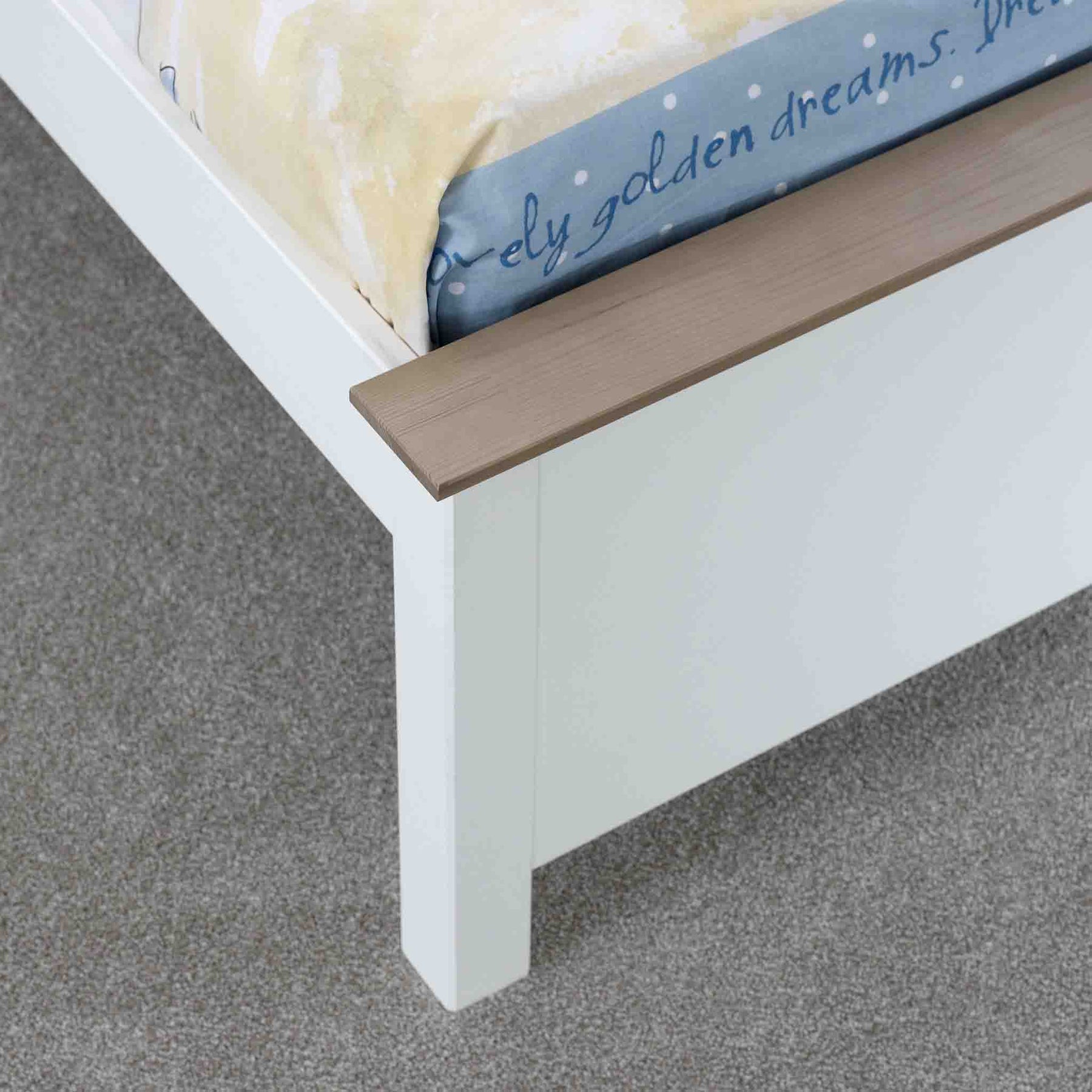 A partially visible cream-colored mattress rests on a white bed frame with a wooden slat, set against a grey carpeted floor.