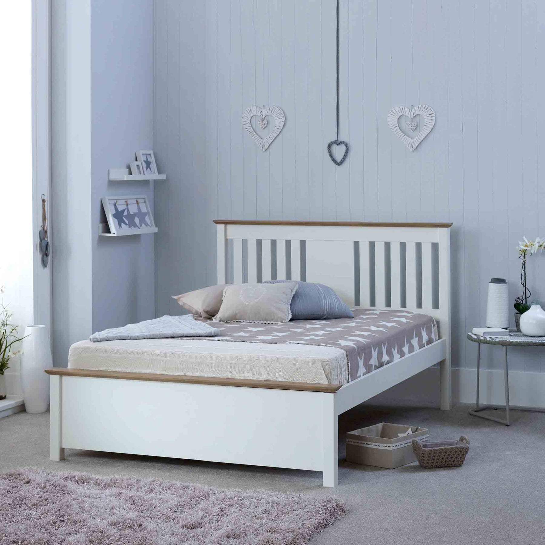 A white, wooden bed with neutral bedding is staged in a clean, gray-walled room adorned with heart ornaments, displaying a serene and tidy bedroom setting.
