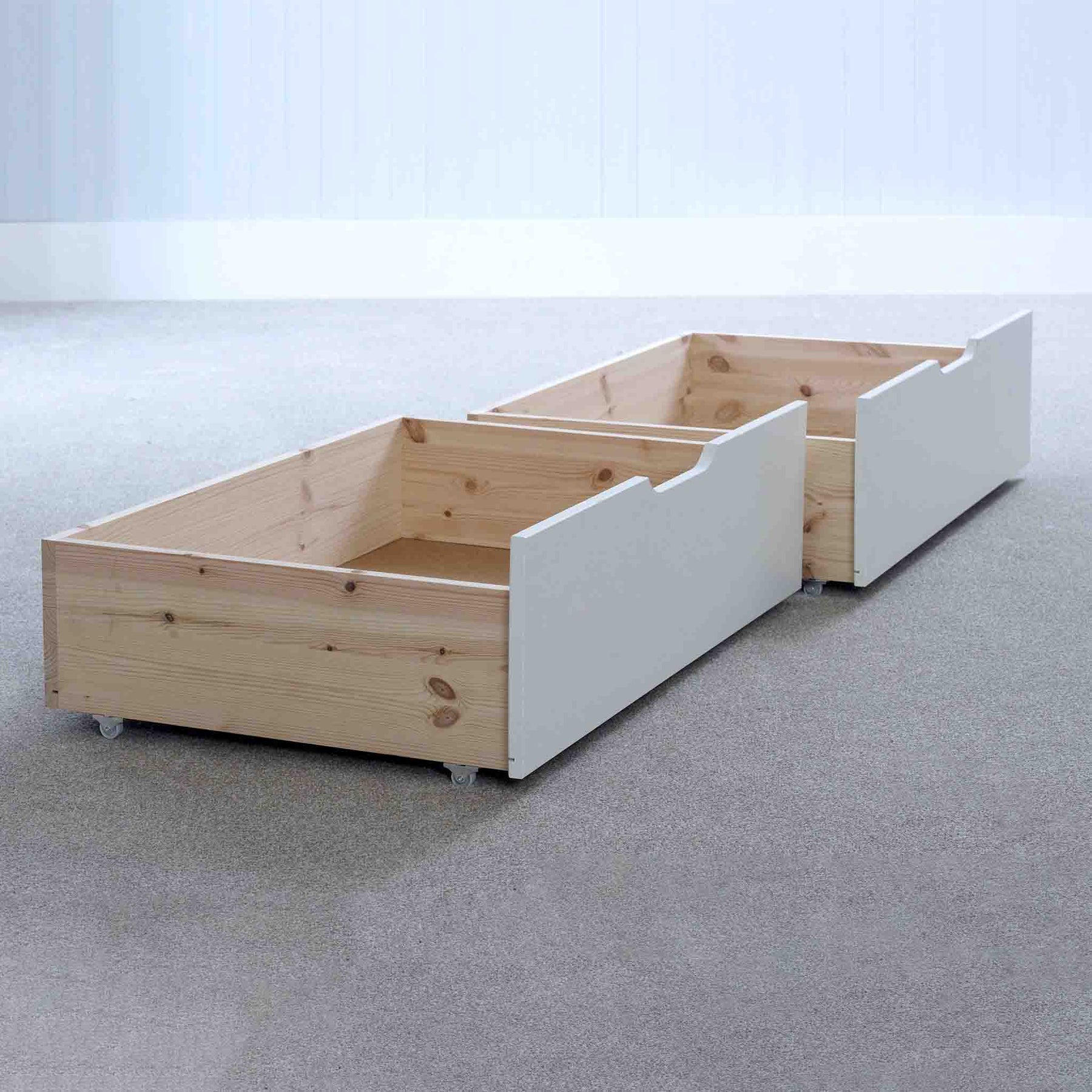 Two wooden under-bed storage drawers on wheels are placed on a light gray carpet; the back drawer has white fronts, providing contrast against the natural wood.