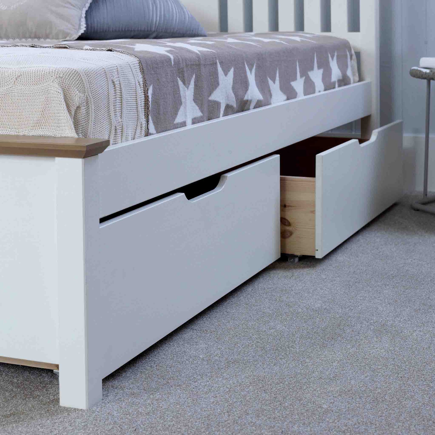 A white bed frame with one drawer partially open, revealing wooden contents, set in a room with a grey carpet and neutral-toned bedding.