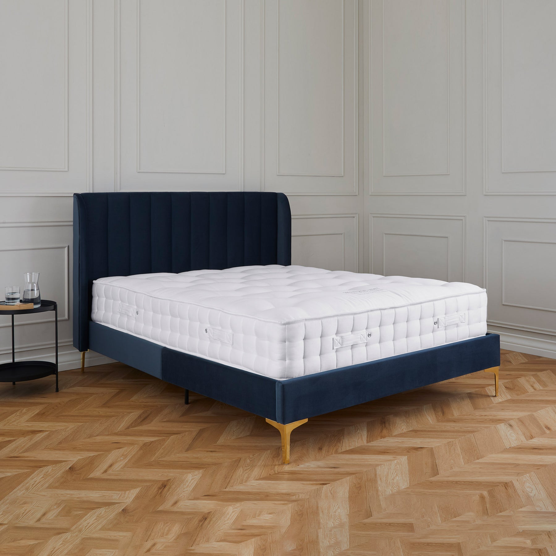 A navy blue upholstered bed with a white mattress is centered in a room with a herringbone wood floor and paneled walls. No text is present.