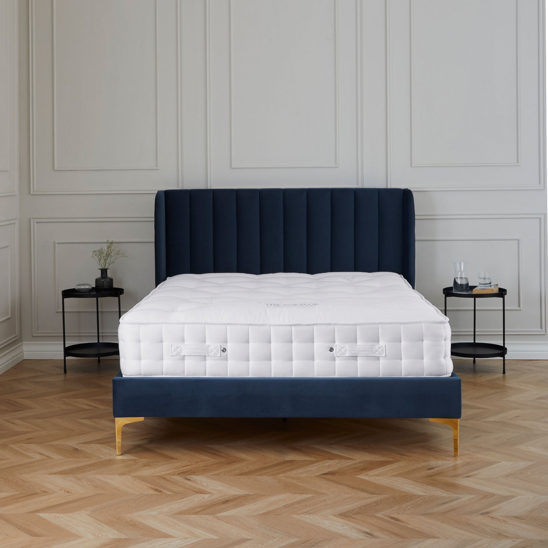 A neatly made bed with a white mattress is placed against a tall blue headboard, set in a room with classic paneled walls and herringbone wood flooring.